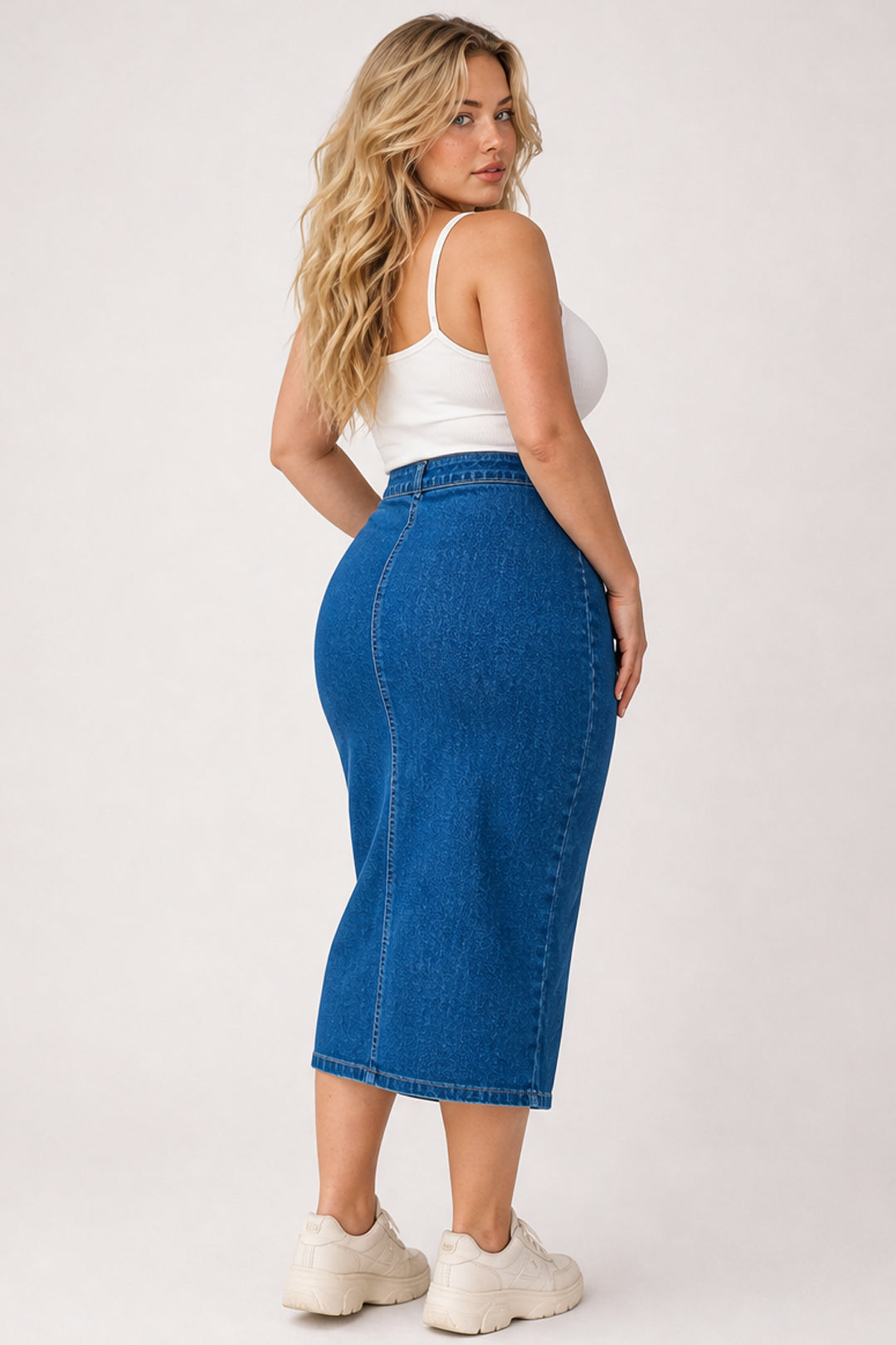 A woman modeling a high-waisted, blue denim midi skirt with a white tank top and white sneakers, photographed from a three-quarters back angle against a light gray studio background.