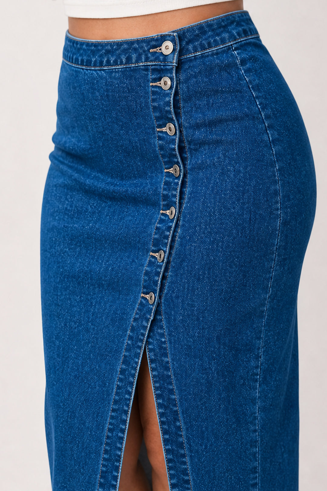 A close-up of a blue denim skirt with a high waist, button-front closure, and a front slit, shown on a model against a plain light background.