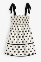 A sleeveless smocked dress with black ribbon shoulder ties, featuring an off-white body with a repeating black bow print and a black horizontal trim line near the hem.