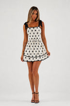 A woman modeling a sleeveless white mini dress with a black repeating bow pattern and black shoulder ties, paired with black strappy heels, against a light gray studio background.