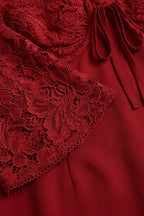 Close-up of a deep red dress featuring a floral lace overlay on the bodice and sleeve, with a matching fabric tie detail and smooth red satin-like fabric underneath.