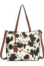 A cow-print tote bag with brown leather handles and a matching hanging tag, shown against a plain white background.