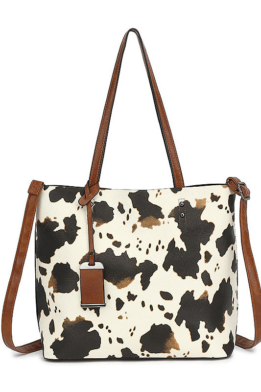 A cow-print tote bag with brown leather handles and a matching hanging tag, shown against a plain white background.