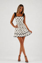 A woman modeling a short white dress with a black bow and bow pattern, black shoulder straps, and black trim, paired with black high-heeled sandals against a light gray studio background.