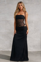 A woman modeling a black strapless maxi dress with a sheer lace corset-style bodice and a flowing satin-like skirt, standing against a textured light gray wall.