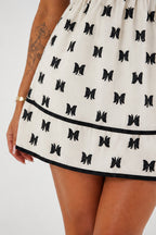 Close-up of a person wearing a white or off-white mini dress with an allover black bow print and a black horizontal trim band near the hem.