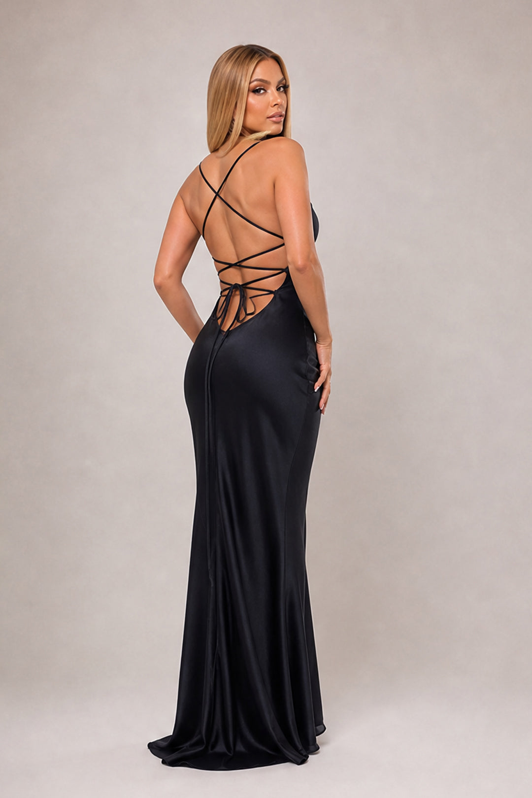 A woman modeling a floor-length black satin gown from the back, showing a strappy open back and a fitted silhouette, against a plain light gray studio background.