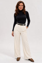 A woman modeling a black long-sleeve mock neck top and high-waisted off-white wide-leg trousers with a thin black belt, paired with leopard print shoes, against a light gray studio background.