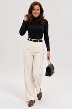 A woman modeling a black long-sleeve mock neck top, cream wide-leg trousers with a thin black belt, leopard-print pointed-toe flats, and carrying a small black handbag with a gold chain strap, against a light gray studio background.