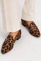 A pair of pointed-toe loafers with a brown and black leopard print and gold-tone horsebit detailing, shown on a model wearing off-white trousers against a light gray studio background.