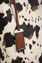A brown leather luggage tag with a matching leather strap, featuring a white plastic insert and black metal trim, laid flat on a black and white cow-print background.