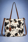 A cow-print tote bag with brown leather handles and a matching hanging tag, set against a blue gradient background.