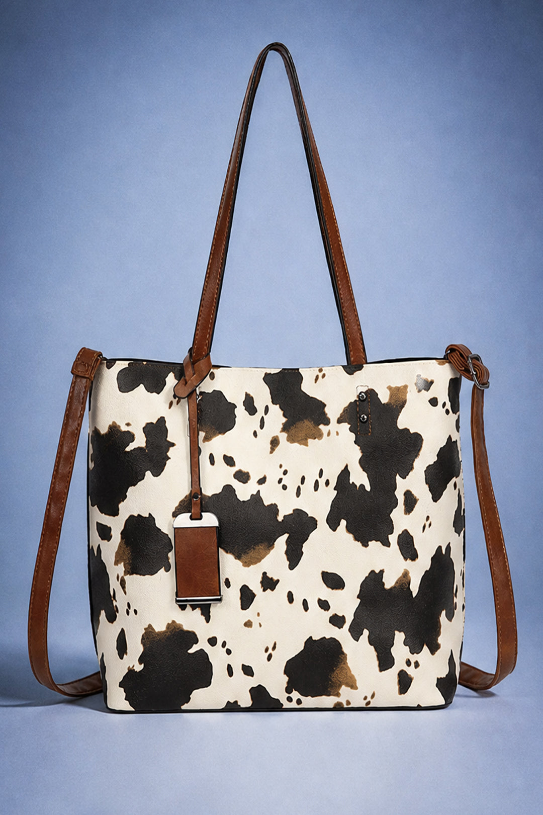 A cow-print tote bag with brown leather handles and a matching hanging tag, set against a blue gradient background.