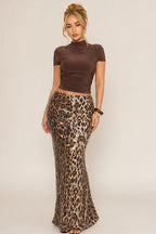 A woman modeling a two-piece outfit consisting of a brown short-sleeve mock-neck crop top and a high-waisted leopard-print pencil skirt with sequins, set against a plain light beige studio background.