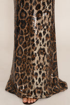 A close-up of a leopard print sequin dress or skirt with a flared hem, worn with brown flat sandals against a light beige studio background.