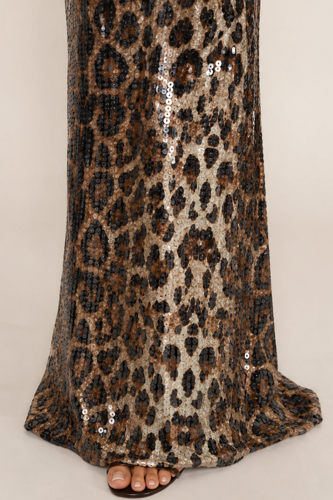 A close-up of a leopard print sequin dress or skirt with a flared hem, worn with brown flat sandals against a light beige studio background.