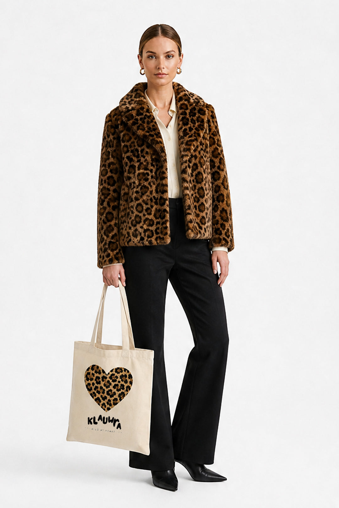 A model stands against a light gray studio background wearing a brown and black leopard-print faux fur jacket over a cream blouse and black trousers, while holding a cream canvas tote bag with a leopard-print heart and the brand name KLAUWRA.