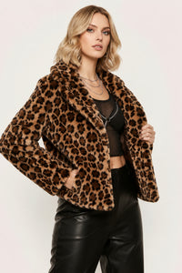 A woman modeling a plush faux-fur leopard print jacket over a black crop top and black faux-leather pants, photographed against a plain light background.