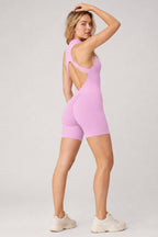 A woman modeling a light pink athletic romper with a back cutout, shown from the back against a light gray studio background.