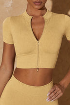 A woman wearing a ribbed short-sleeve crop top with a front zipper and a matching high-waisted ribbed skirt in a warm mustard yellow color, set against a textured beige background.