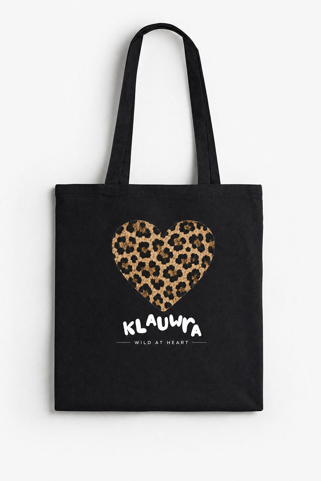 A black canvas tote bag with long black handles, featuring a leopard-print heart graphic and the white text 'KLAUWRA WILD AT HEART' on the front, shown against a plain white background.