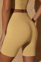A close-up of a model wearing ribbed high-waisted biker shorts in a warm mustard yellow color, paired with a matching ribbed crop top, set against a textured beige stone wall.