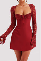 A woman wearing a red mini dress with long lace sleeves, a sweetheart neckline, and a small front bow, shown against a light gray studio background.