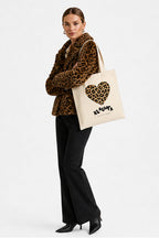 A model wearing a leopard-print jacket and black pants carries a cream canvas tote bag with a leopard-print heart and the brand name 'KLAUWRA' printed on it, set against a plain white studio background.