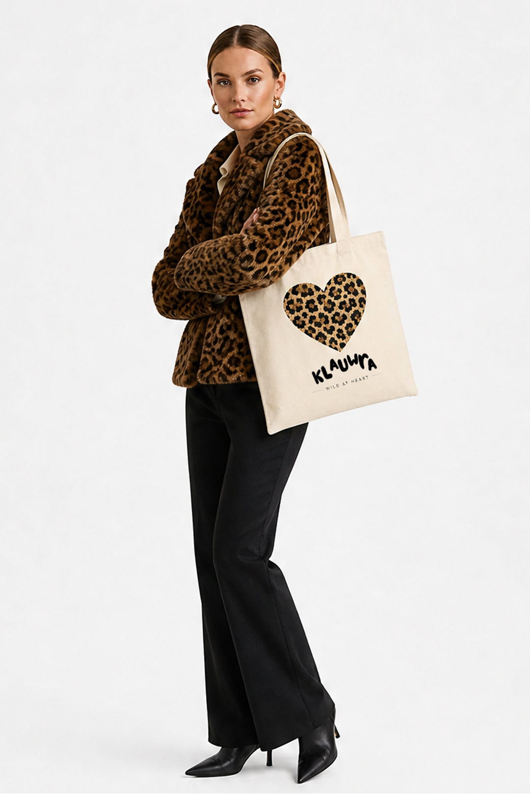 A model wearing a leopard-print jacket and black pants carries a cream canvas tote bag with a leopard-print heart and the brand name 'KLAUWRA' printed on it, set against a plain white studio background.