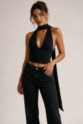 A woman modeling a black halter crop top with a long draped tie detail and black high-waisted jeans against a plain light gray studio background.