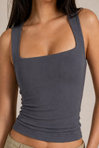 A close-up of a model wearing a dark gray ribbed tank top with a square neckline and wide shoulder straps, paired with light beige pants against a neutral beige background.