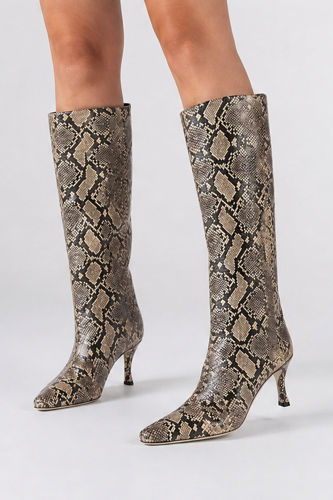 A pair of knee-high pointed-toe heeled boots with a snakeskin pattern in beige, brown, and gray, shown on a model's legs against a light gray studio background.