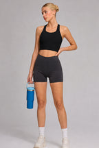 A woman modeling a black sports bra and dark gray athletic biker shorts, holding a blue water bottle against a solid gray studio background.