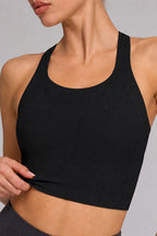 A close-up of a woman wearing a black ribbed athletic sports bra with a scoop neckline and wide shoulder straps, shown against a light gray studio background.