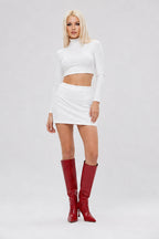 A blonde model wearing a white long-sleeve crop top and matching white mini skirt, paired with shiny red knee-high boots, against a light gray studio background.
