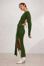 A woman models a ribbed knit two-piece set consisting of a long-sleeve top and matching high-slit skirt in olive green, styled with white ankle boots against a neutral studio backdrop.