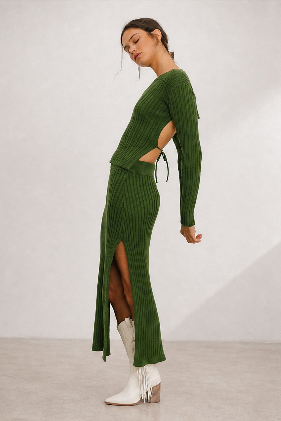 A woman models a ribbed knit two-piece set consisting of a long-sleeve top and matching high-slit skirt in olive green, styled with white ankle boots against a neutral studio backdrop.