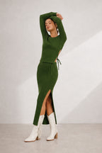 A model wearing a ribbed knit green midi dress with a thigh-high slit and a matching drawstring belt, paired with white fringed ankle boots, set against a neutral studio backdrop.