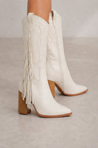 A pair of off-white leather cowboy boots with fringe detailing and a block heel, shown on a model's feet against a neutral gray background.