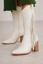 White faux leather western-style boots with fringe detailing and a stacked wood-look block heel, shown on a model wearing a green ribbed skirt.