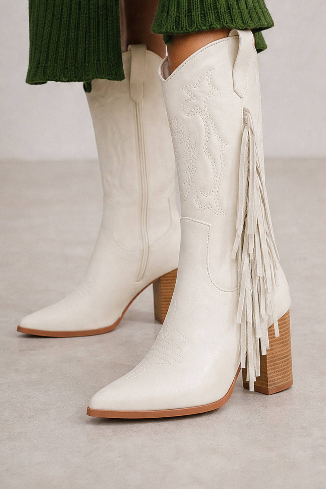 White faux leather western-style boots with fringe detailing and a stacked wood-look block heel, shown on a model wearing a green ribbed skirt.