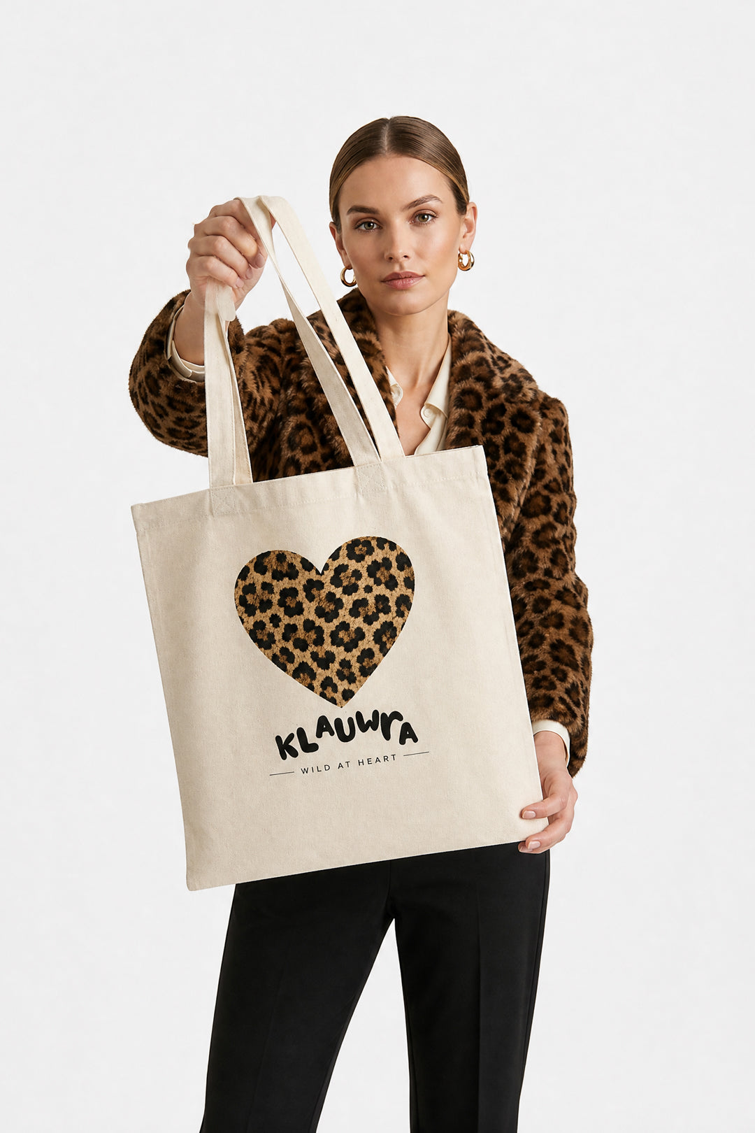 A woman wearing a leopard-print jacket holds up a cream canvas tote bag with a leopard-print heart graphic and the brand name 'KLAUWRA' printed below it, against a plain light gray background.