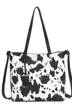 A black and white cow print tote bag with black shoulder straps and a black hanging tag, shown against a plain white background.