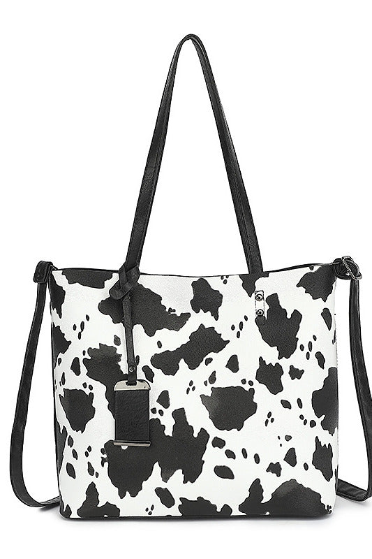 A black and white cow print tote bag with black shoulder straps and a black hanging tag, shown against a plain white background.