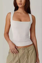 A woman wearing a fitted white square-neck tank top and beige pants, cropped from the mouth down against a plain light gray studio background.
