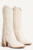 A pair of off-white faux leather cowboy boots with fringe detailing on the outer side and a low block heel, shown against a plain white background.