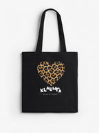A black canvas tote bag with two shoulder straps, featuring a leopard-print heart graphic and the text 'KLAUWRA WILD AT HEART' in white on the front, set against a plain white background.