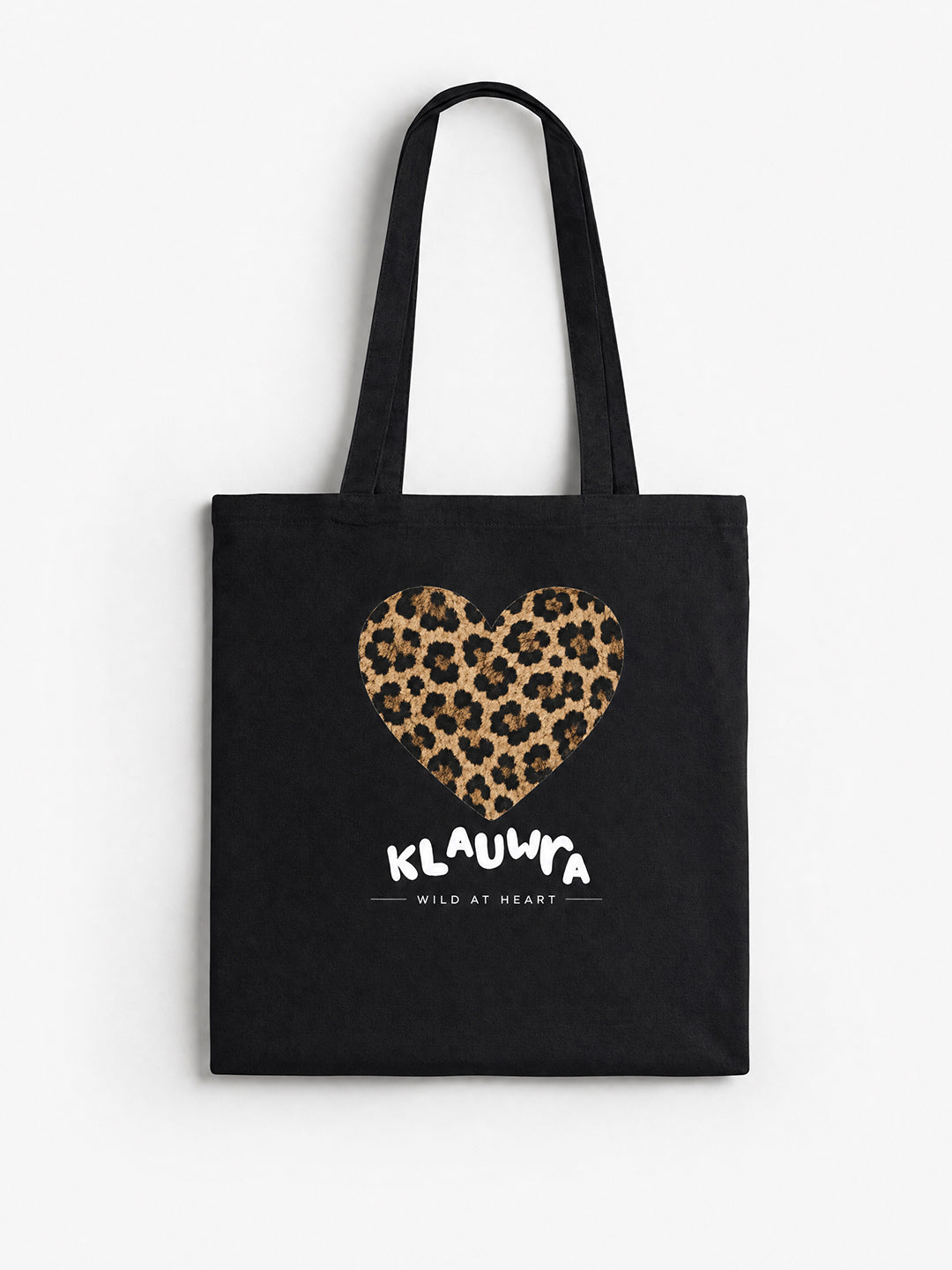 A black canvas tote bag with two shoulder straps, featuring a leopard-print heart graphic and the text 'KLAUWRA WILD AT HEART' in white on the front, set against a plain white background.