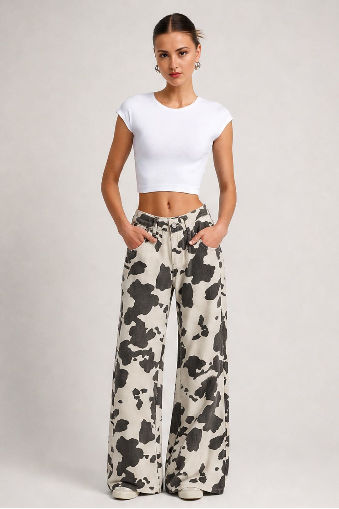 A woman modeling a white short-sleeve crop top and black-and-white cow-print wide-leg pants against a light gray studio background.