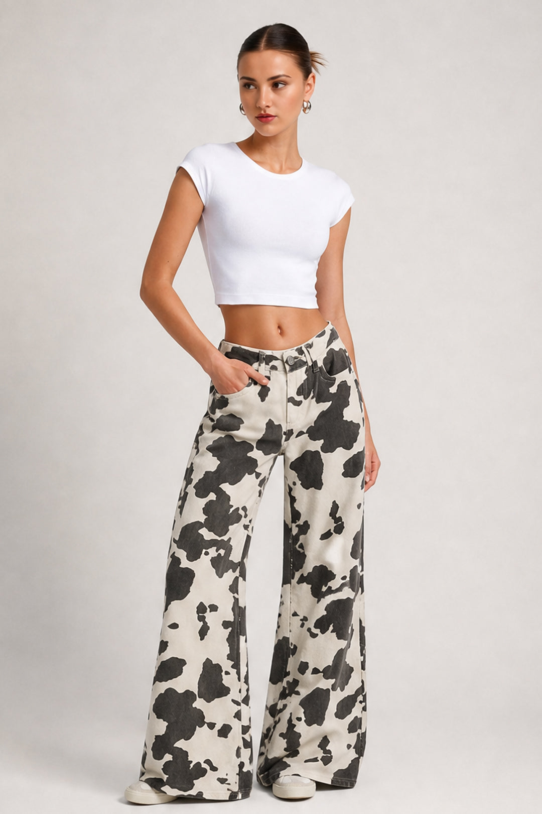 A woman modeling a white cropped short-sleeve top and high-waisted, wide-leg pants with a black and off-white cow print, standing against a light gray studio background.